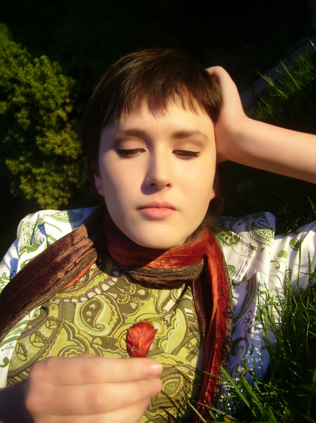 Image: a person with short hair, a green shirt, and a red scarf  stares at a leaf in their hand