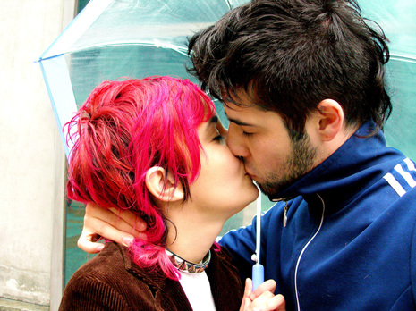 Image: a person with pink hair wearing a studded collar and holding an umbrella kisses another person with a blue jacket