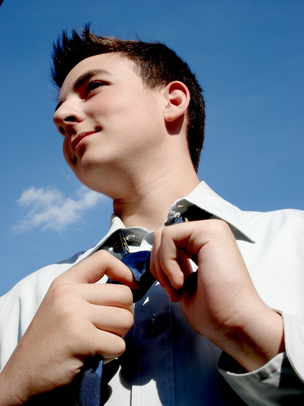 Image: a person stands against a blue sky, tying a blue necktie around their neck